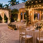 Outdoor wedding reception with warm string lights, white draped arch, and floral decorations along a lantern-lit aisle.