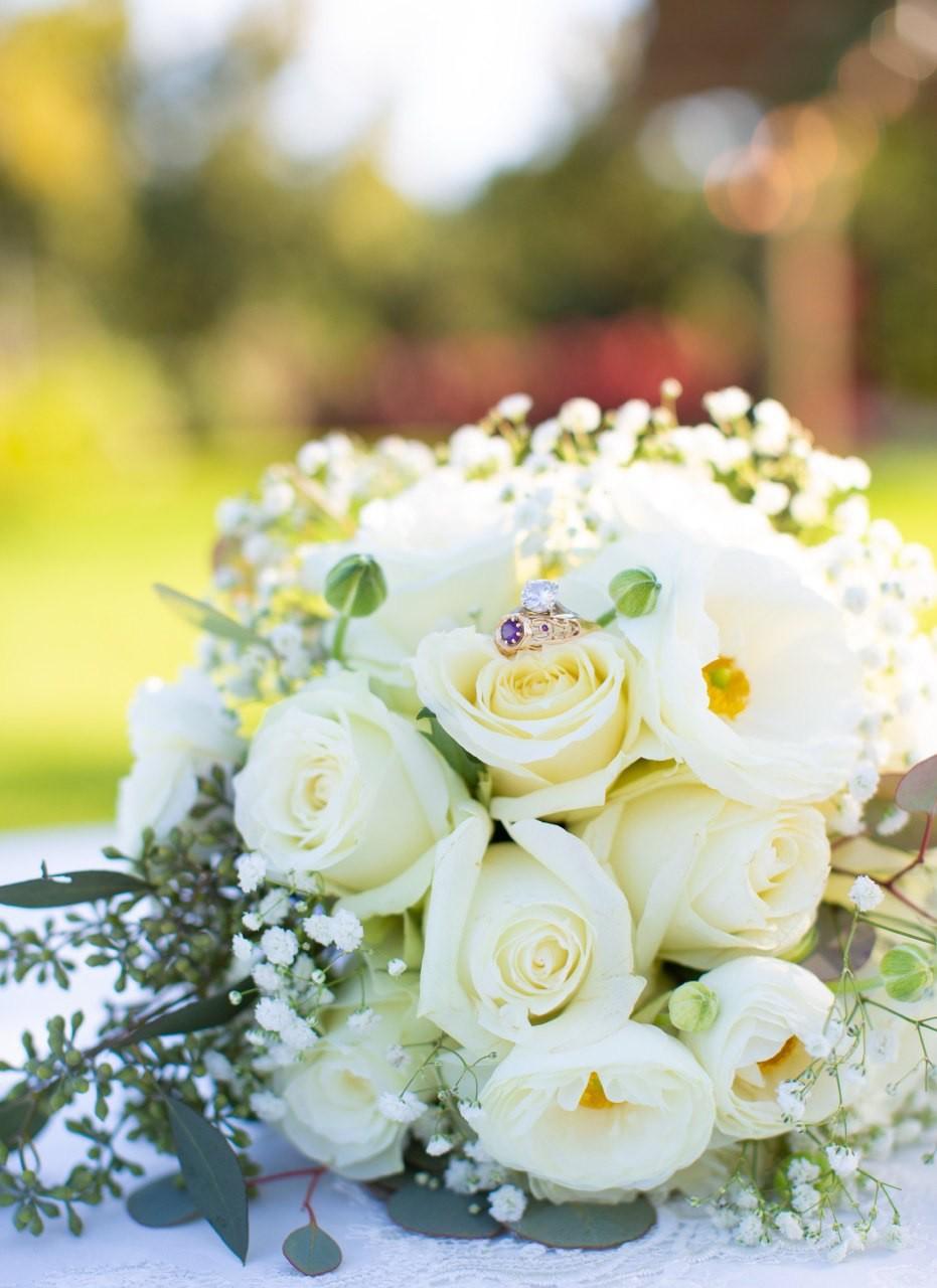 Bouquet of white roses with baby's breath; a diamond ring with a purple gemstone rests on a bloom.