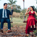 Couple in formal attire swinging on a wooden swing set in a park, smiling at each other; man in a navy suit and tie, woman in a red dress.